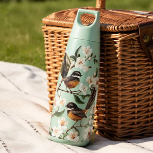 100%NZ Chatty Fantails Drink Bottle next to a brown basket on a picnic blanket.