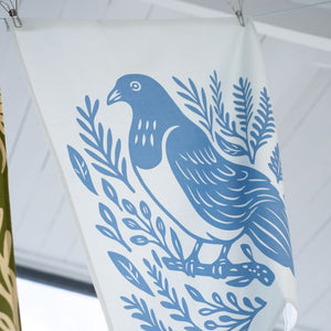 100%NZ Woodcut Kererū cotton tea towel hanging from a line shown a blue kererū with foliage.
