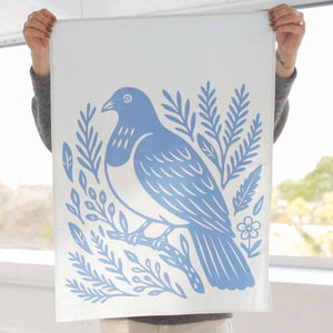 Person holding the 100%NZ Woodcut Kererū Tea Towel with a blue bird design against a blurred outdoor background.