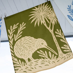 100%NZ Woodcut Kiwi Tea Towel pegged to a line, shown hanging outside.