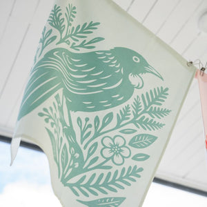 100%NZ Woodcut Tūī Tea Towel pegged to a line shown hanging outside.