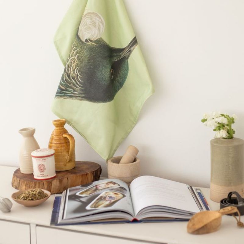 100%NZ Hushed Green Tūī Tea Towel hanging in a kitchen next to an open cookbook.