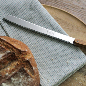 André Verdier Laguiole Debutant Bread Knife – Country, resting diagonally on a light blue-gray striped kitchen towel next to a loaf of bread.