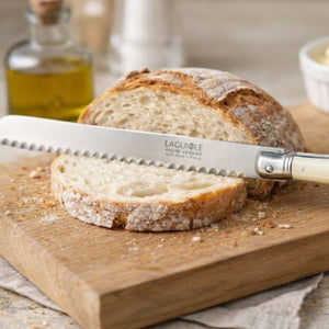André Verdier Laguiole Debutant Bread Knife in Ivory resting on a wooden cutting board with sliced bread, and a bottle of olive oil blurred in the background.