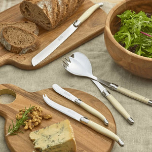 A set of cream-colored and stainless steel serving utensils, including a cheese knife, bread knife, fork, and server by André Verdier, styled on a linen tablecloth with wooden boards.