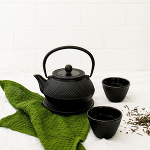 Avanti Hobnail Cast Iron Teapot next to two small black cups, a green textured cloth, and scattered loose tea leaves, with a white tiled wall in the background.