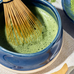 A close-up of a blue ceramic bowl filled with green matcha tea, with a bamboo whisk from the Avanti Matcha Ceremonial Set resting inside the bowl on a textured fabric surface.