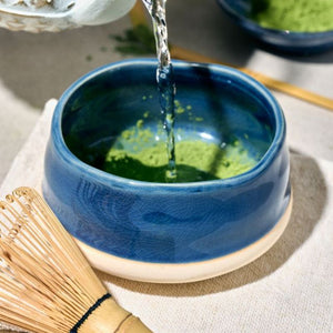 A blue ceramic matcha whisk bowl with a beige base from the Avanti Matcha Ceremonial Set, shown with hot water being poured into it and green matcha powder inside. A bamboo whisk and another bowl are partially visible in a styled setting.