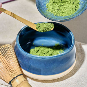 A blue ceramic matcha bowl with a beige base, filled with green matcha powder, alongside a bamboo whisk and spoon from the Avanti Matcha Ceremonial Set.