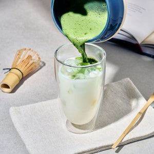 Green matcha tea being poured from a blue ceramic pitcher into a double-walled glass of iced milk, styled on a light gray surface with a bamboo whisk, book, and napkin.