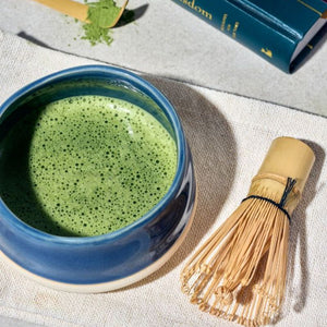 A blue ceramic bowl from the Avanti Matcha Ceremonial Set, filled with frothy green matcha tea sits on a textured linen surface, alongside a bamboo whisk and a small spoon of loose matcha powder.