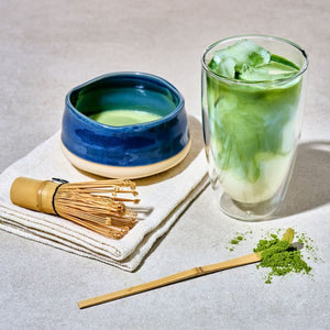 A styled matcha tea setup on a light gray surface, featuring a double-walled glass of green matcha iced with milk, a blue ceramic bowl of matcha, and a bamboo whisk from the Avanti Matcha Ceremonial Set.