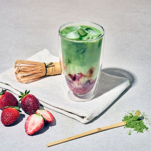 A green drink in a clear double-walled glass with ice, styled on a folded white cloth with strawberries, next to the Avanti Matcha Whisk and Scoop Set on a light grey surface.