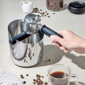 Avanti Stainless Steel Knock Box styled next to coffee beans, a cup of coffee and a person holding a portafilter on a grey background.