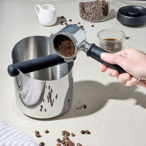 Avanti Stainless Steel Knock Box styled next to coffee beans, a cup of coffee and a person holding a portafilter.