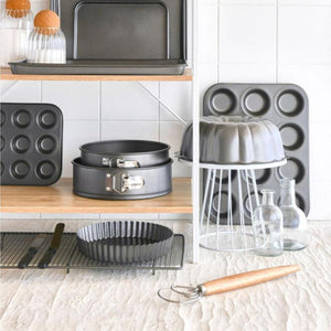 Collection of baking pans and tools on a kitchen counter, including the Bakemaster Dough Whisk, with a white tiled wall background.