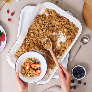 Baked granola being served onto the Bakemaster Enamel Cereal Bowl with berries on a beige surface.