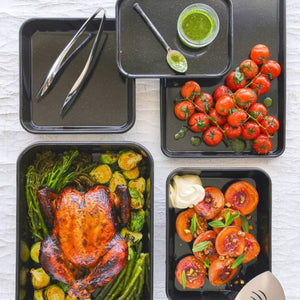 Bakemaster Professional Vitreous Enamel collection styled with roasted vegetables and roasted chicken, with utensils and a small container of green sauce.