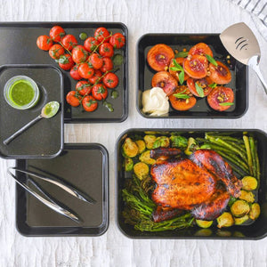 Bakemaster Professional Vitreous Enamel collection styled with roasted vegetables and roasted chicken, with utensils on a light wooden surface.