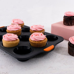Bakemaster Silicone Muffin Pan – 12 Cup, shown filled with cupcakes on a light gray surface. The background includes a pink block holding another cupcake.