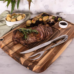 Carved roast beef served on an acacia carving board with bread, vegetables, and carving knives.