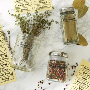 Glass jars containing dried herbs and spices with labels on a marble surface.