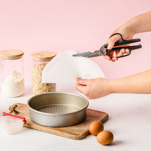 Person cutting a piece of paper with the Dreamfarm Bishears in the kitchen scissors position on a kitchen counter with baking ingredients.