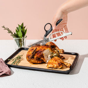 Roasted chicken on a cutting board with a pair of Dreamfarm Bishears in kitchen shears position and a glass of herbs on a light pink background.