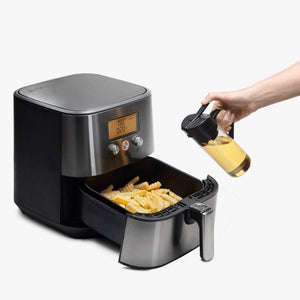 Person holding the Dreamfarm Oileroo next to an air fryer filled with fries on a white background.