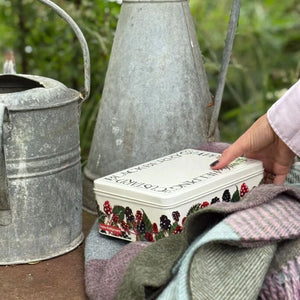 Person holding Emma Bridgewater Blackberries Biscuit Tin in a rustic outdoor scene with watering cans and wool blanket nearby.