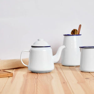 Falcon Enamel Teapot - White with Blue Rim on a wooden table next to an enamel utensil holder.