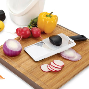 OXO Good Grips Hand Held Mandoline Slicer on a chopping board with sliced onions, radishes, and vegetables.