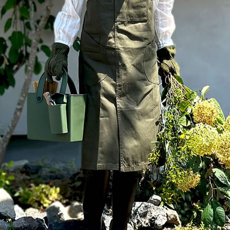 Woman wearing green apron holding flower cuttings in one hand and the Hachiman Garden Tool Bag with Pocket in the other - the pocket shown attached to the front of the bag
