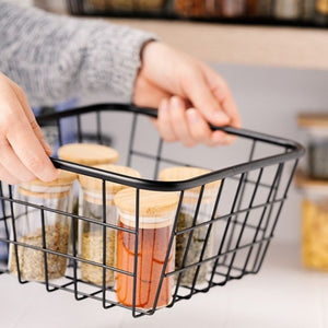 Person holding a black wire basket with spice jars in a kitchen setting.