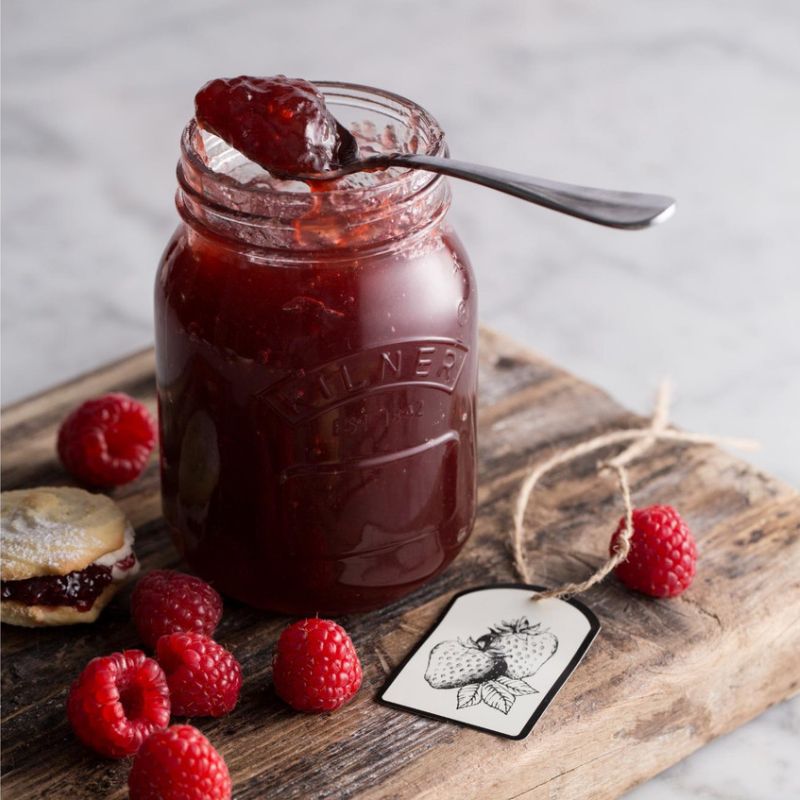 A 500mL Kilner Genuine Preserve Jar filled with raspberry jam on a rustic wooden chopping board