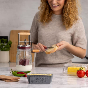 Woman spreading butter on bread using the Kilner butter spreader, with the Kilner butter churner, butter dish and silicone butter moulds in the foreground.
