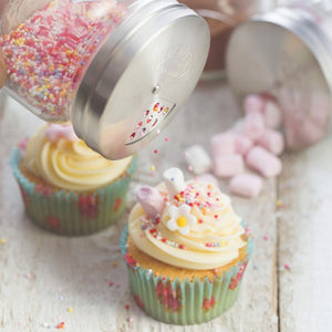 Sprinkles being shaken from Kilner shaker jar onto cupcakes topped with icing and marshmallows.