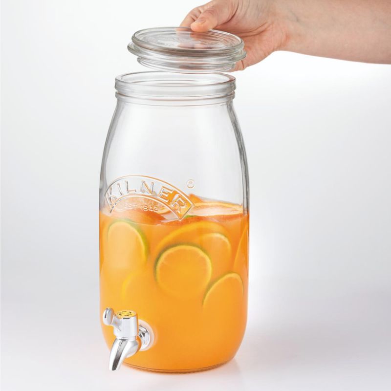 Clear glass drink dispenser by Kilner filled with orange juice and lime slices, with a hand holding the lid.
