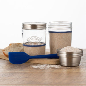 Kilner glass jars with metal lids, a blue spatula, and measuring cup on a wooden surface.