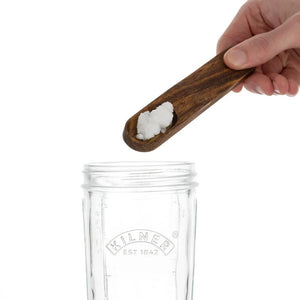 Hand holding a wooden scoop with kefir grains over a clear Kilner jar on a white background