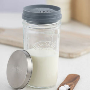 A Kilner glass jar with a gray strainer lid, shown with its stainless steel metal lid leaning beside it on a light surface. The jar is partially filled with a kefir milk, and a small wooden spoon with kefir grains sits in the foreground.