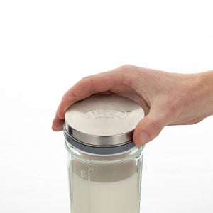 A hand holding a stainless steel lid on a clear kilner glass jar with a grey seal, shown against a plain white background.