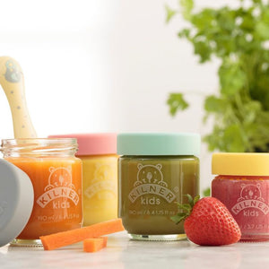 Four Kilner Kids jars with different colored lids on a white surface with a blurred green plant in the background.