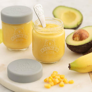 Two jars of yellow baby food in the 190 mL Kilner Kids Jars with a spoon, surrounded by avocado, banana, and corn on a white surface.