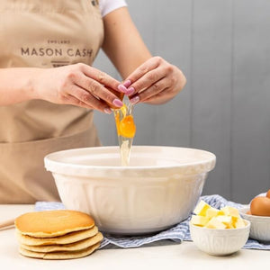 Egg being cracked into the Mason Cash Cream Colour Mixing Bowl on a table with pancakes and butter