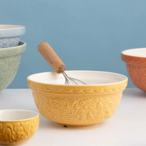 A yellow embossed ceramic mixing bowl with a white interior by Mason Cash, shown with a wooden-handled whisk resting inside. Other ceramic bowls in different colours are partially visible in the background against a soft blue wall.