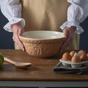 Person holding the Mason Cash In The Forest Bear Mixing Bowl in Cane colour  on a wooden table with eggs and a wooden spoon.