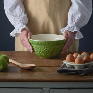 Person holding the Mason Cash In The Forest Green Hedgehog Mixing Bowl on a wooden table with eggs and apples.