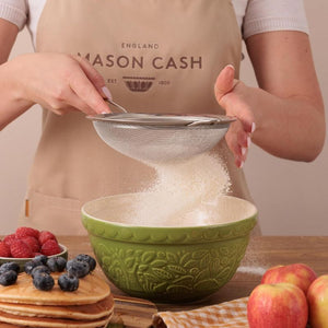 Person sifting flour into the Mason Cash In The Forest Green Hedgehog Mixing Bowl with fruits and pancakes on a table