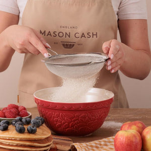 Person sifting flour into the Mason Cash In The Forest Hedgehog Red Mixing Bowl with fruits and pancakes on a table.